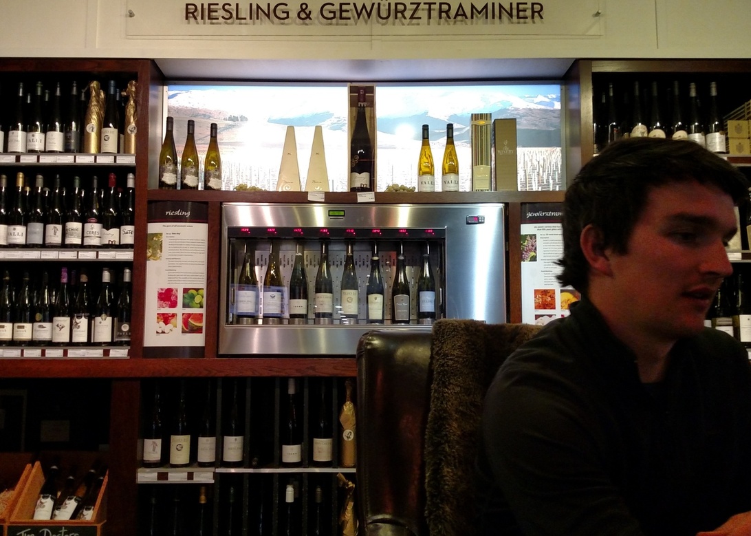 Ein anspruchsvoller Weinverkostungsbereich mit einem Wein Konservierungs- und Ausschanksystem, das Weißweine (Riesling und Gewürztraminer laut Beschilderung) präsentiert. Die Einrichtung umfasst mehrere Flaschen, die auf Holzregalen und in temperaturgesteuerten Spendern ausgestellt sind. Eine Person ist im Vordergrund sichtbar, was darauf hindeutet, dass dies ein Kundenbereich in einer Weinbar oder einem Restaurant ist.